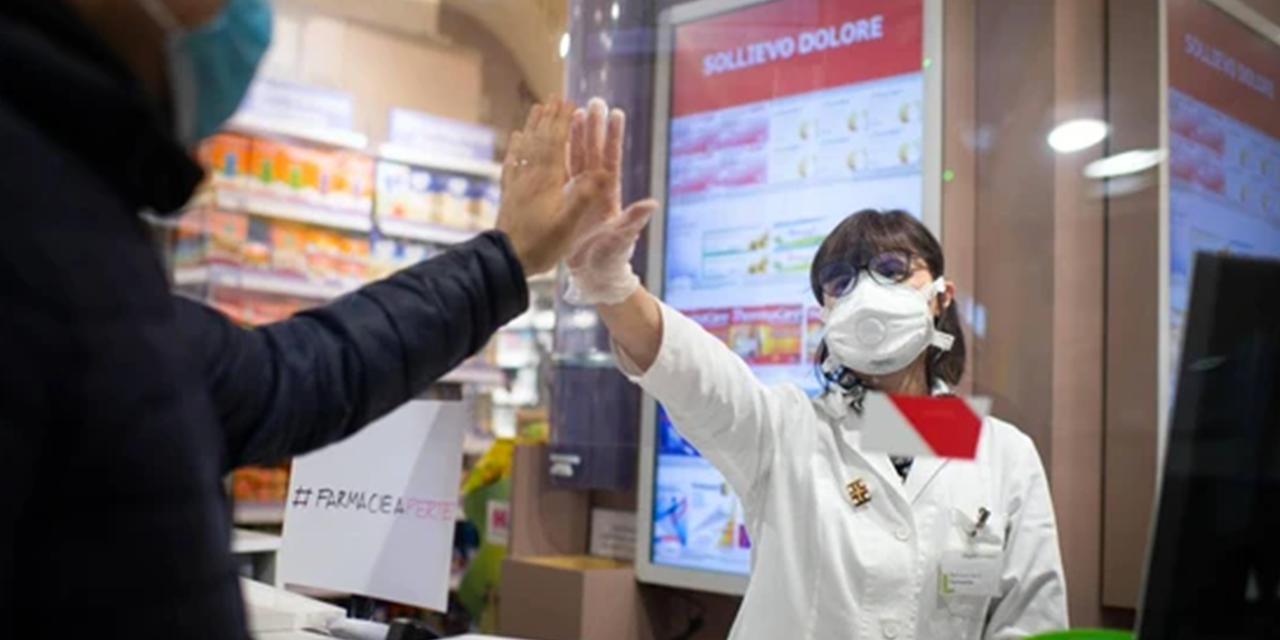una farmacista protetta da un vetro separatore protettivo allunga la mano verso un cliente.